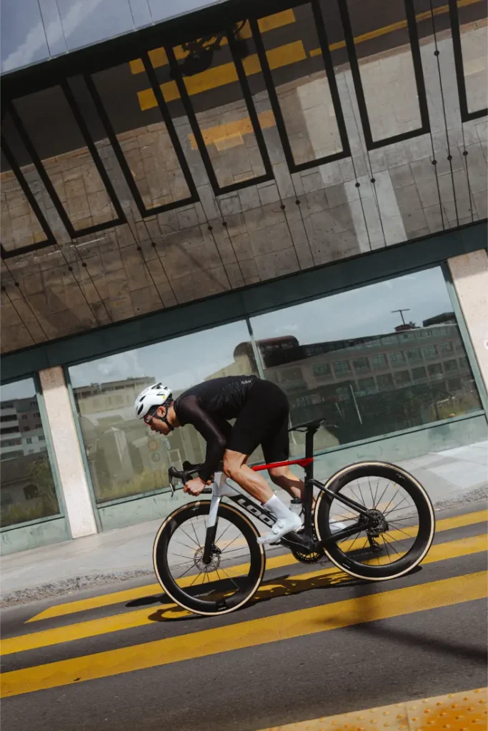 Un ciclista vestido de negro y con casco blanco cruza en su bicicleta de carretera LOOK 795 Blade RS 3 un paso de peatones con rayas amarillas en una zona urbana con edificios de cristal, que recuerda al moderno paisaje urbano de Nevers.