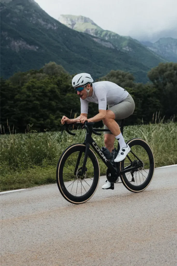 Un ciclista, con casco blanco y equipación de ciclismo, circula en una bicicleta de carretera LOOK 795 Blade RS 3 negra por una carretera asfaltada cerca de Nevers, con montañas verdes y árboles al fondo.