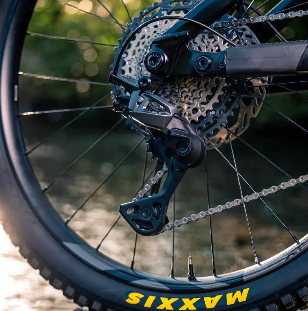 Primer plano de la rueda trasera de una bicicleta de montaña, en el que se aprecian el cambio mecánico Shimano DEORE XT M8200, el cassette con todo detalle, la cadena y el neumático Maxxis, con un fondo al aire libre difuminado.