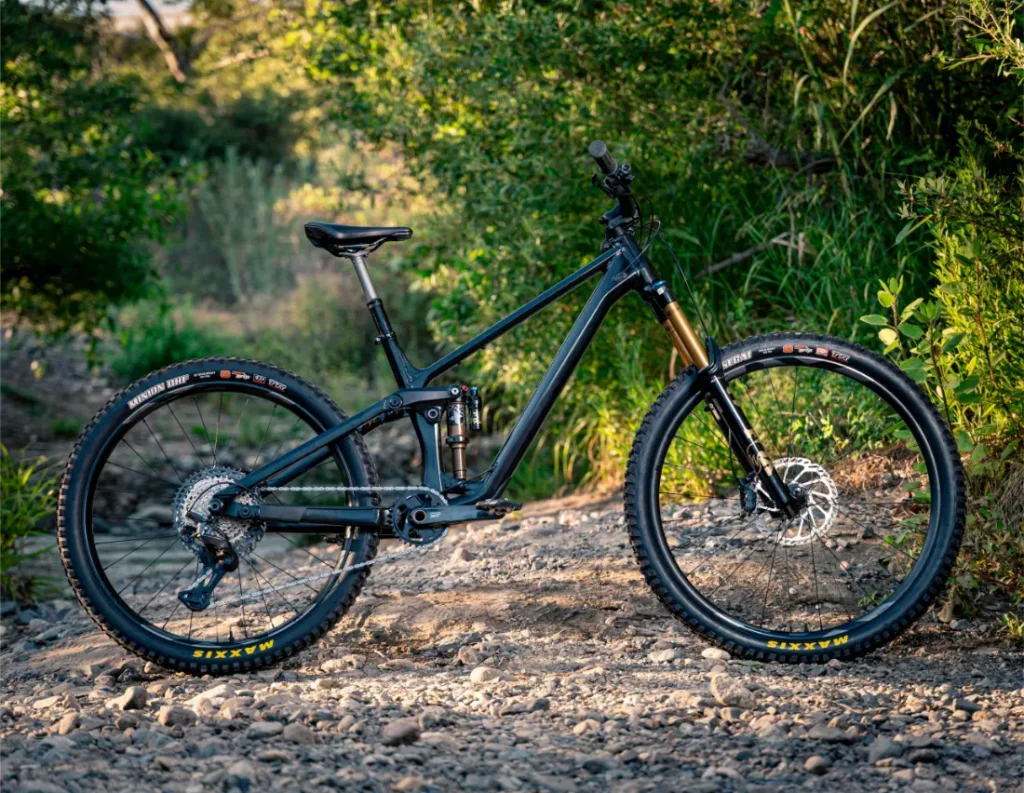Una bicicleta de montaña de doble suspensión con cuadro negro, cambio mecánico Shimano DEORE XT M8200 y neumáticos de tacos se encuentra en un camino de tierra rocoso rodeado de frondosa vegetación. «Larga vida al cable» en cada aventura extrema.