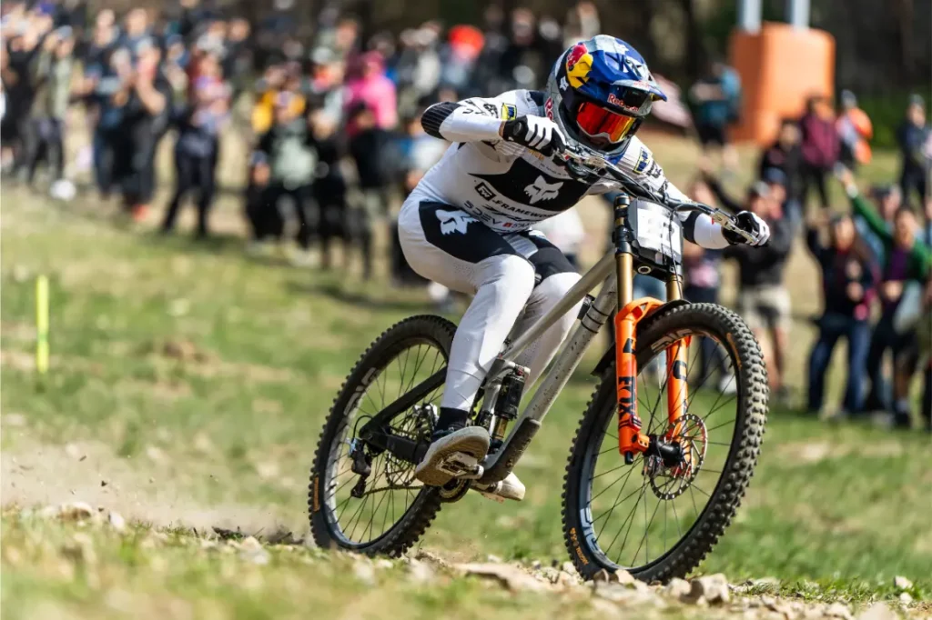 Un ciclista de montaña, vestido con un maillot de competición blanco y negro, desciende a toda velocidad por un sendero rocoso ante una multitud de espectadores, demostrando la precisión del TRP EVO PRO Anniversary Edition en este evento de edición limitada celebrado en la Península Ibérica.