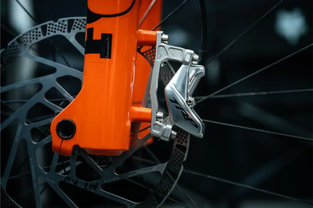 Primer plano de una pinza y un disco de freno delantero TRP EVO PRO Anniversary Edition en una horquilla naranja, en el que se ven los radios y parte de la rueda; esta edición limitada se inspiró en los recorridos por la Península Ibérica.