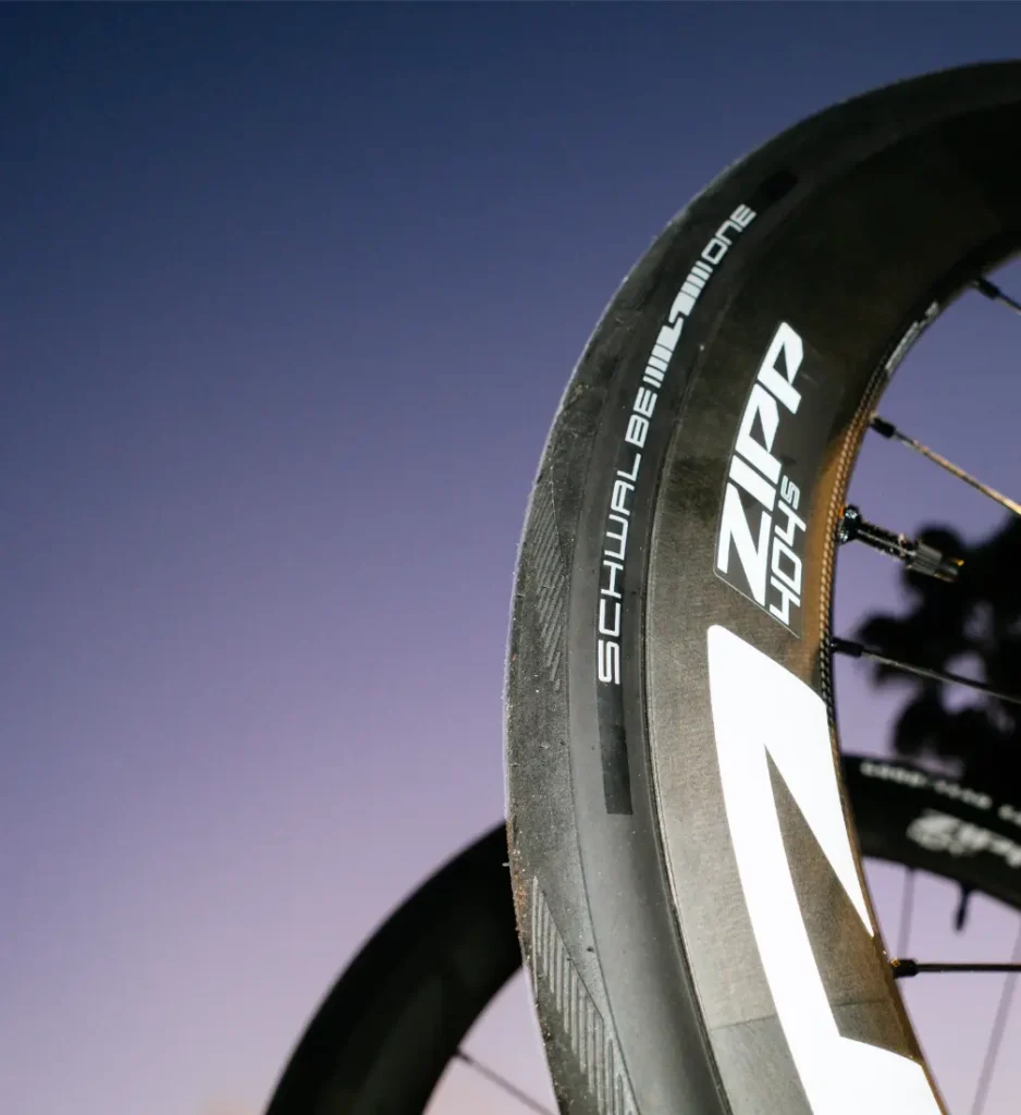 Primer plano de una rueda de bicicleta Zipp 404 S con un neumático Schwalbe One, con un cielo morado claro de fondo, que destaca su velocidad y su calidad superior a un precio impresionante.