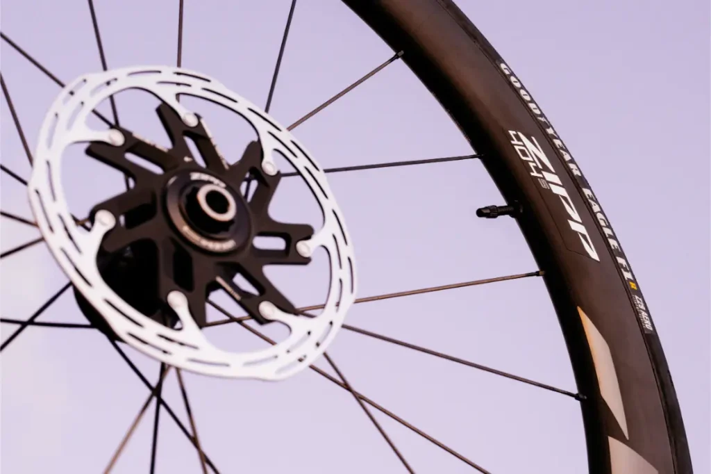 Primer plano de una rueda de bicicleta Zipp 404 S con radios negros, un disco de freno y una llanta con el logotipo de la marca, con un cielo de color morado pálido de fondo; diseñada para la velocidad y el rendimiento.