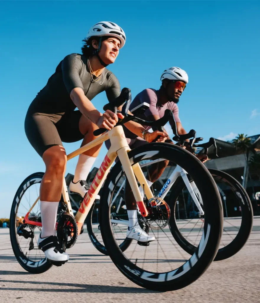 Dos ciclistas, equipados con casco y ropa de ciclismo, pedalean en bicicletas de carretera Zipp 404 S uno al lado del otro en un día soleado, con un cielo azul y despejado de fondo, disfrutando tanto de la velocidad como del rendimiento.