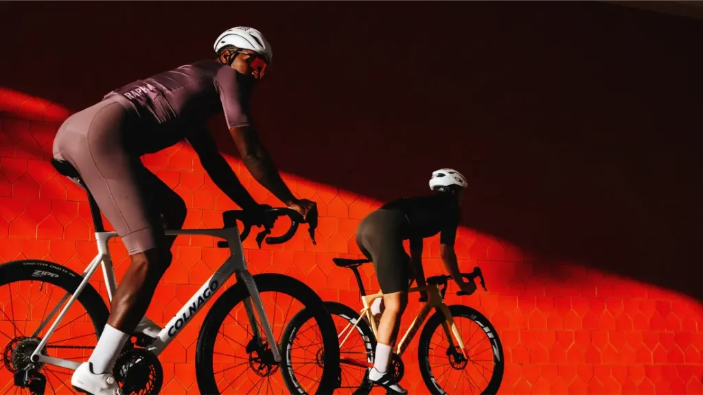 Dos ciclistas, con casco y equipación de ciclismo, pedalean en sus bicicletas de carretera Zipp 404 S junto a una pared de un rojo intenso, iluminados por un espectacular rayo de sol que evoca tanto la velocidad como el estilo.
