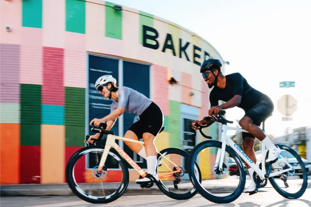 Dos ciclistas, con casco y montados en bicicletas de carretera Zipp 404 S, pasan a toda velocidad junto a un edificio de colores en el que se lee la palabra «BAKERY», haciendo gala de su velocidad.