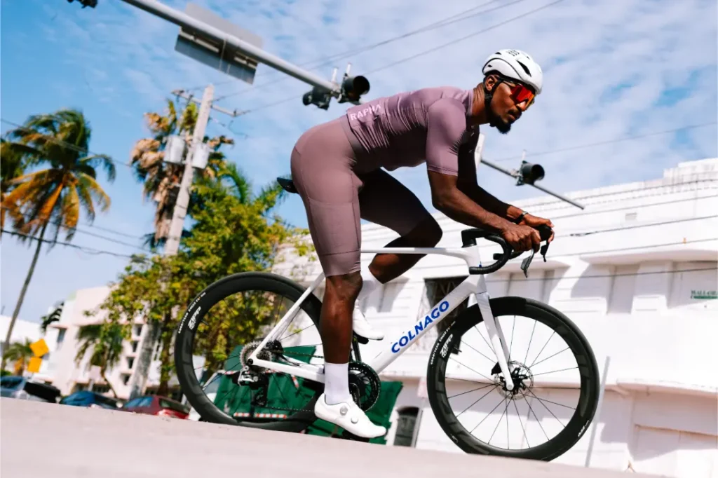 Un ciclista, que lleva casco y gafas de sol, pedalea en una bicicleta de carretera Colnago blanca con ruedas Zipp 404 S, disfrutando de la velocidad por una calle urbana soleada, flanqueada por palmeras y edificios.