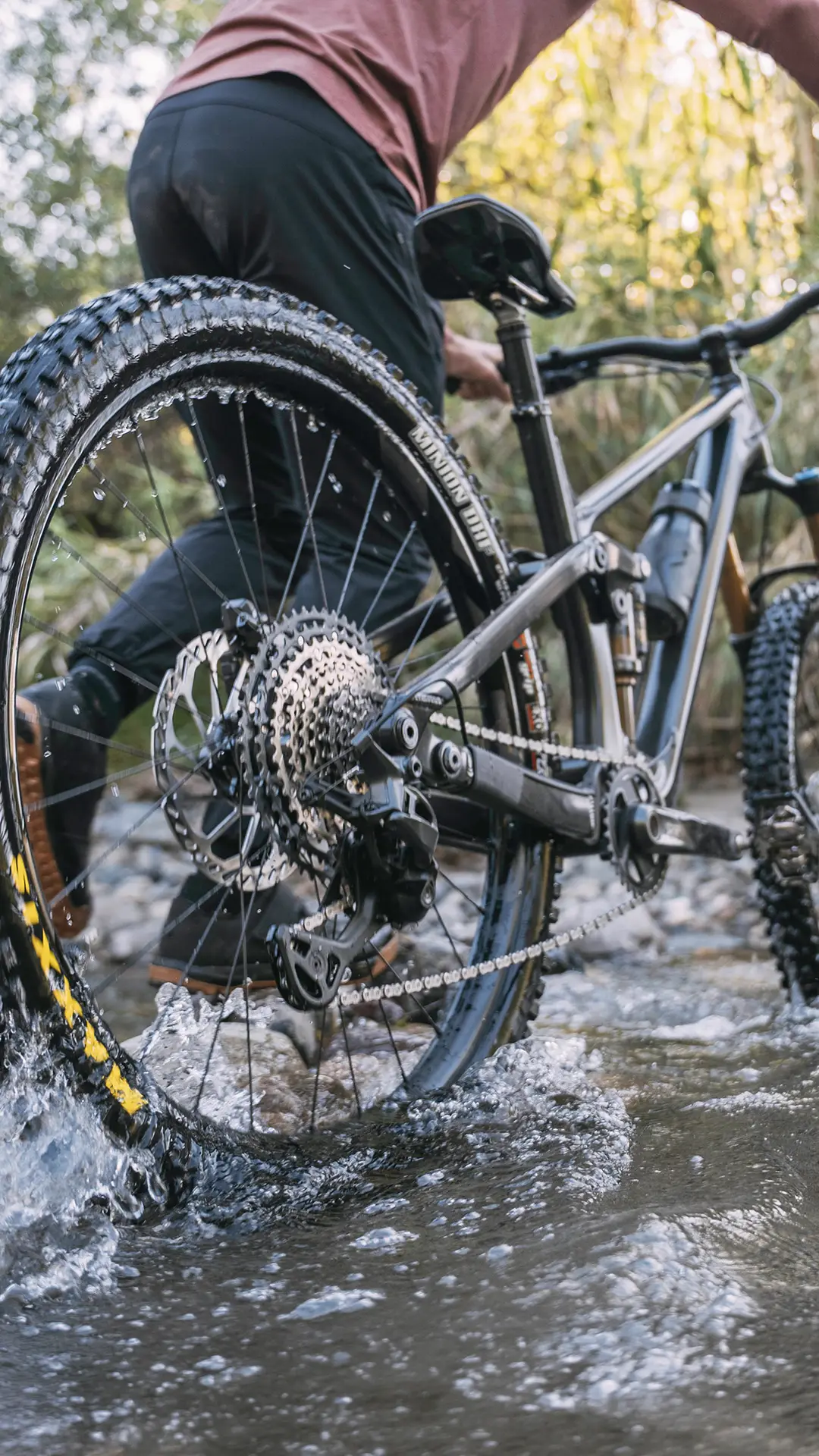 Una persona empuja una bicicleta de montaña equipada con un cambio mecánico Shimano DEORE XT M8200 a través de un arroyo poco profundo, con el agua salpicando alrededor de las ruedas de la bicicleta, lo que garantiza una larga vida útil del cable incluso en condiciones difíciles.