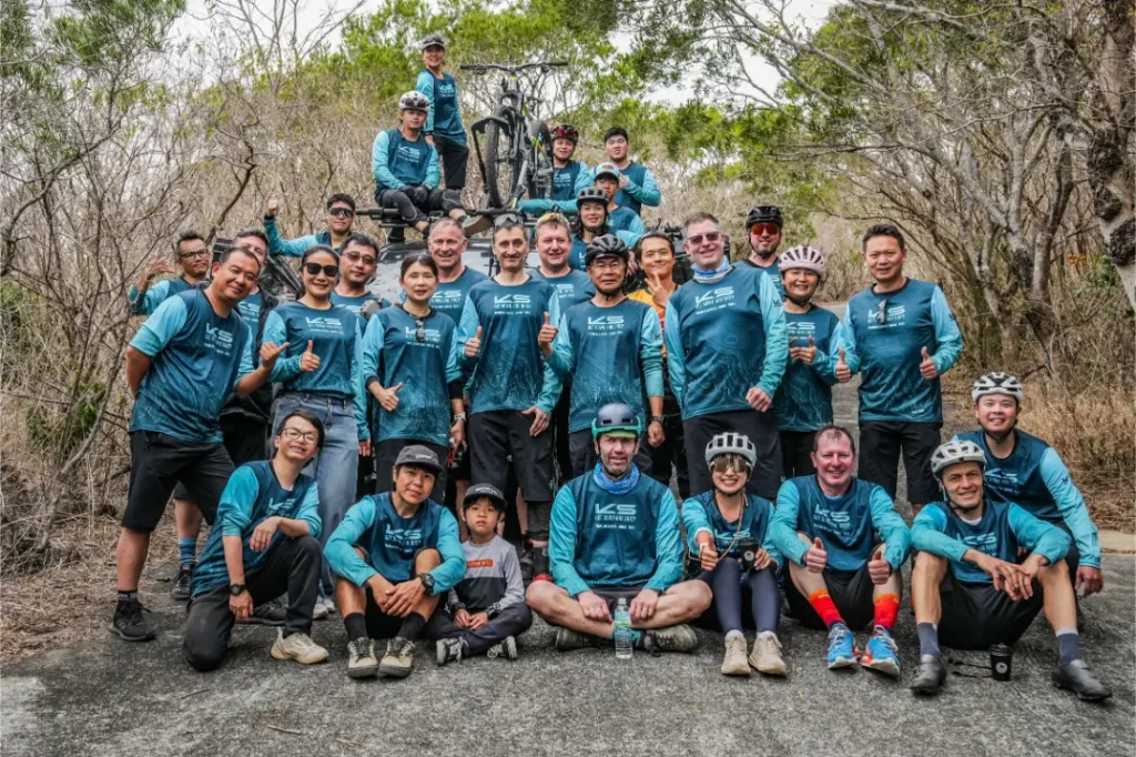Un grupo de ciclistas con maillots azules a juego posan juntos al aire libre, unos de pie y otros sentados, rodeados de árboles y vegetación seca, representando con orgullo a KS Sportmed y su distribución oficial.