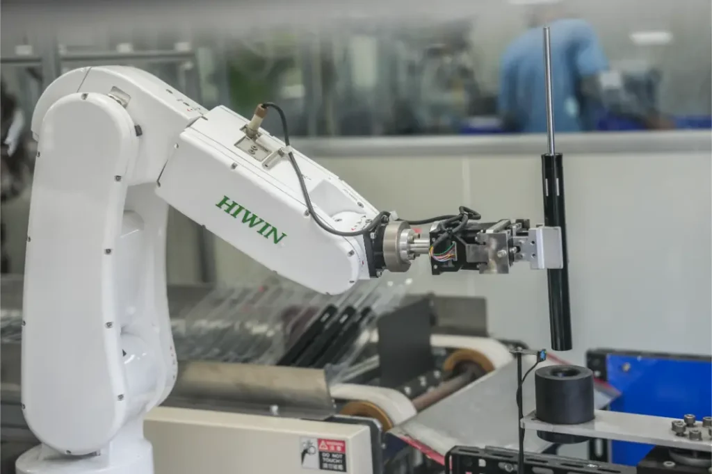 Un brazo robótico HIWIN sujeta un objeto cilíndrico en un entorno de fabricación o laboratorio, con KS como distribución oficial en España; al fondo se ve maquinaria y una persona.