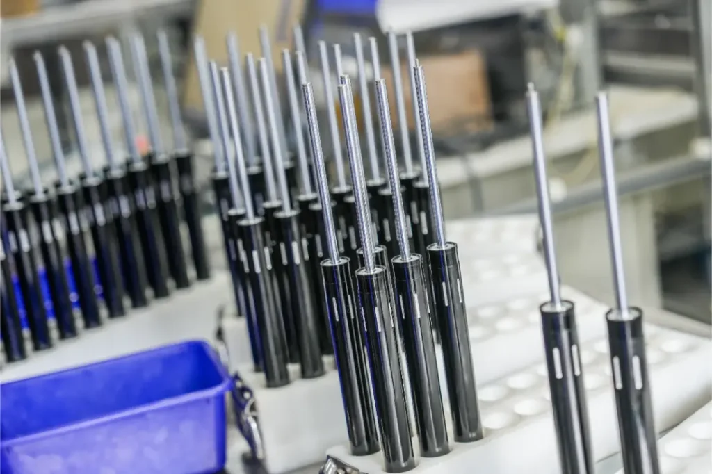 Filas de componentes metálicos y negros parcialmente ensamblados, posiblemente amortiguadores o cilindros neumáticos, se alinean en un entorno de fábrica o taller con contenedores azules cerca, destacando la distribución oficial KS España de Sportmed.