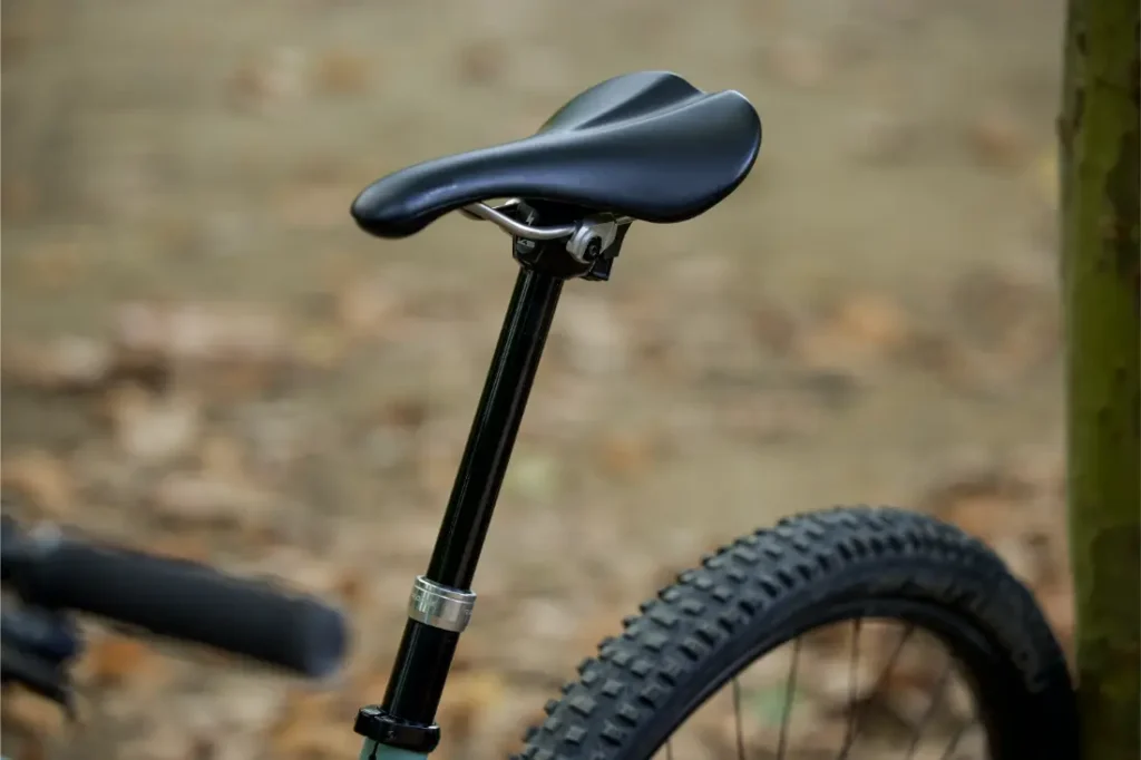 Primer plano de un sillín y una tija de bicicleta KS negros con un neumático trasero de tacos, tomados al aire libre cerca de un árbol con un fondo borroso y terroso. Distribución oficial Sportmed.