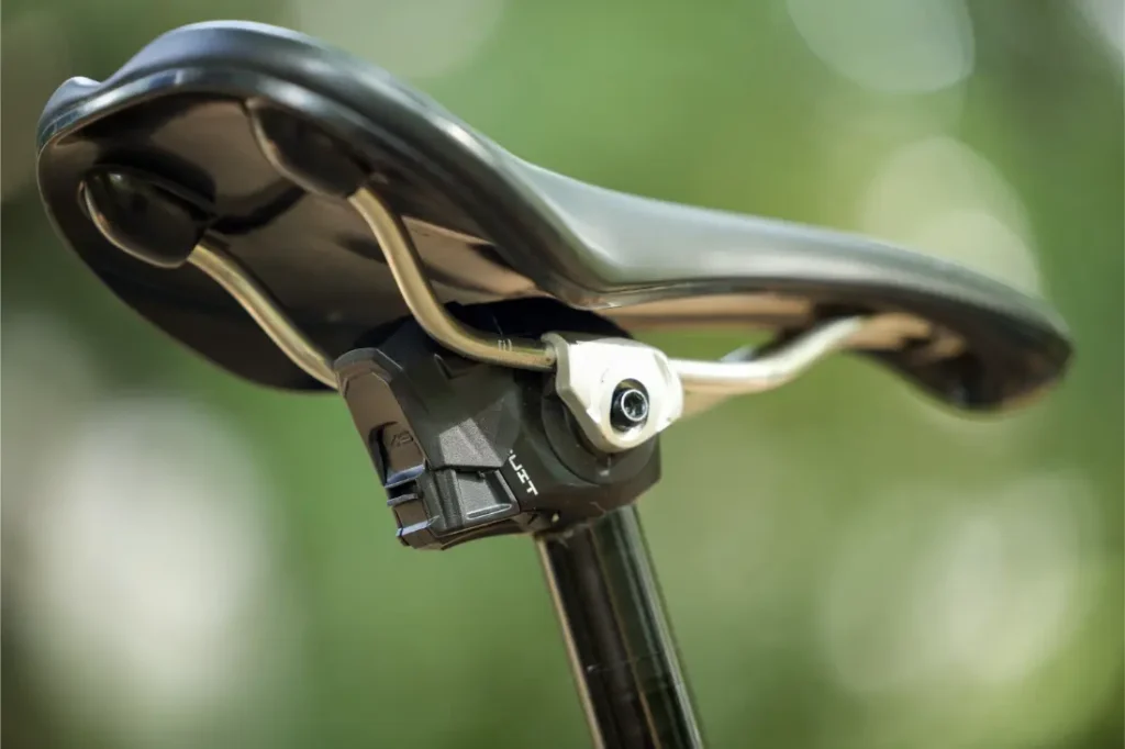 Primer plano de un sillín de bicicleta sujeto a una tija de sillín, con un componente electrónico debajo del sillín. La imagen se refiere a la distribución KS de Sportmed en España, sobre un fondo verde difuminado.