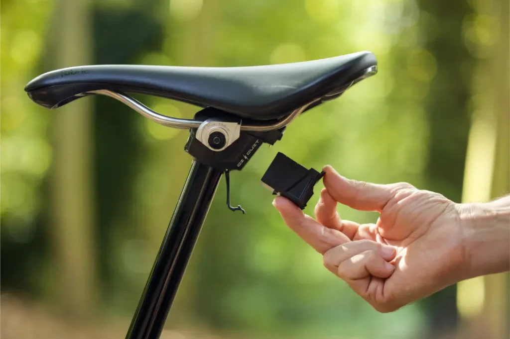 Una mano inserta un pequeño dispositivo negro bajo el sillín de una bicicleta, con un fondo verde borroso al aire libre, destacando Sportmed y la distribución oficial KS España.