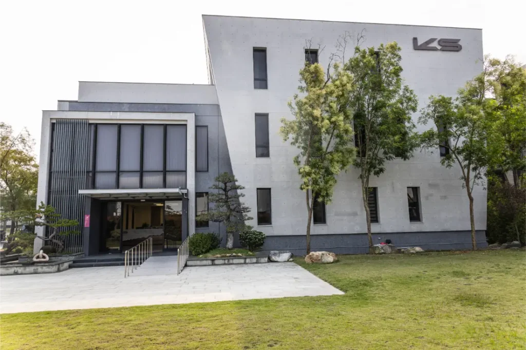 Moderno edificio de dos plantas con fachada gris, grandes ventanales y las letras "KS" en la parte superior derecha, sede de la distribución oficial Sportmed. Los frondosos árboles y el césped proporcionan un primer plano acogedor.