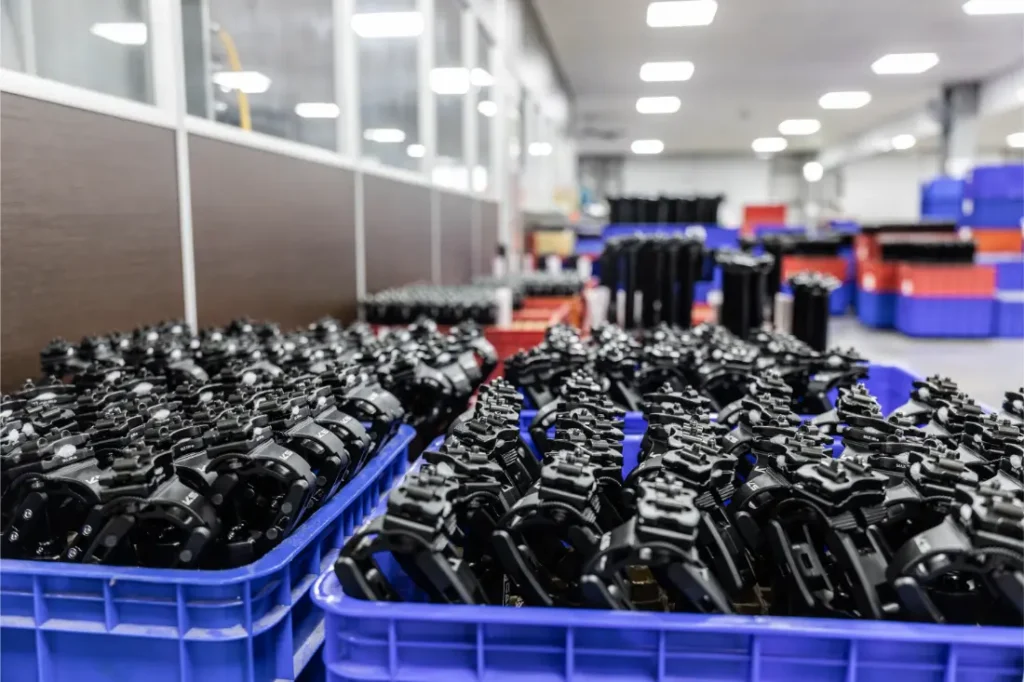 Filas de cajas azules llenas de piezas metálicas negras se organizan en el interior de unas instalaciones industriales bien iluminadas, destacando la distribución oficial de KS dentro del proceso de producción.