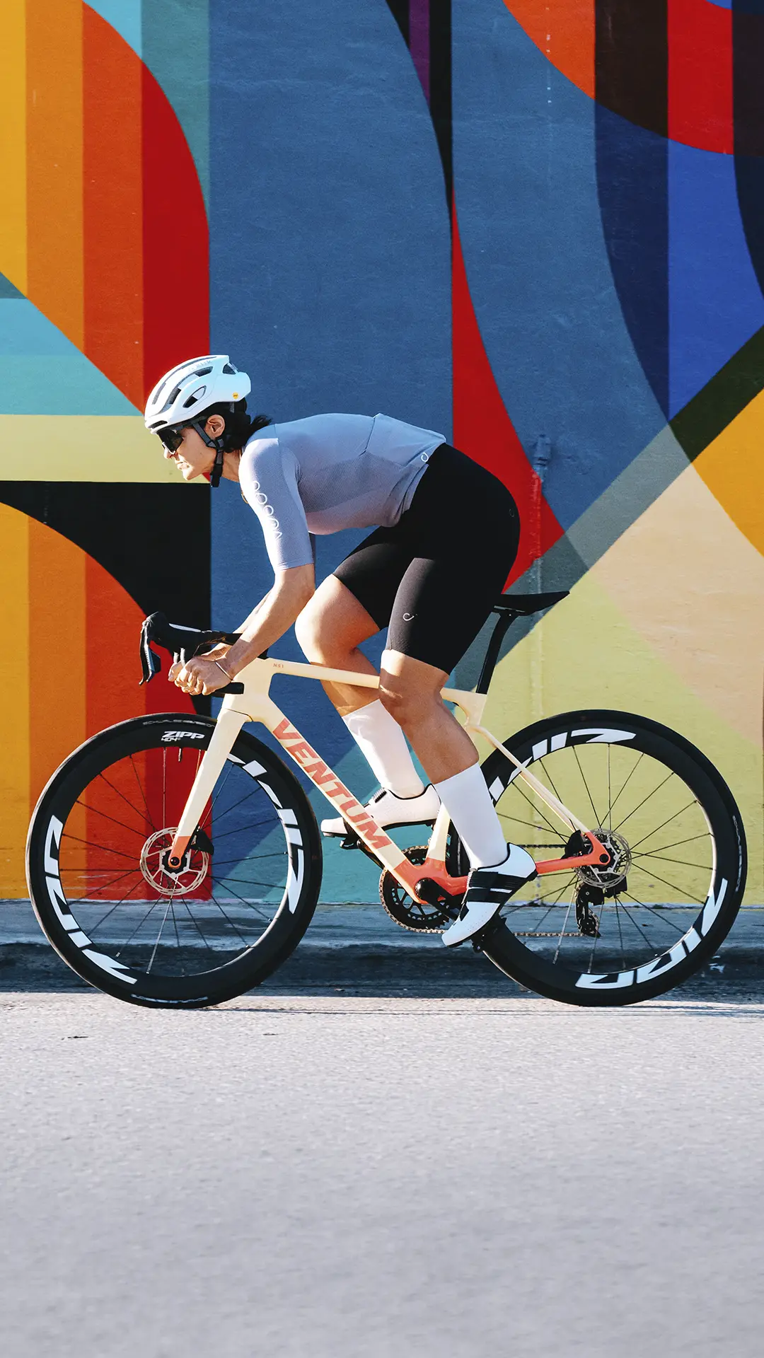 Un ciclista, que lleva casco y gafas de sol, pasa a gran velocidad junto a un colorido mural geométrico montado en una bicicleta de carretera blanca equipada con ruedas Zipp 404 S.
