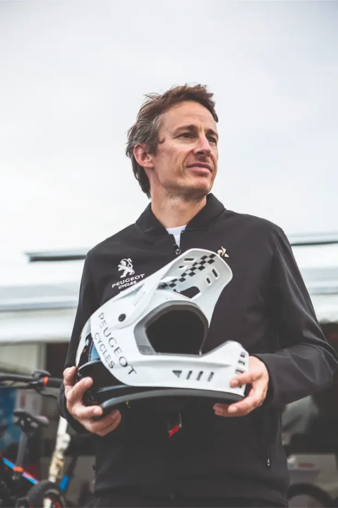 Un hombre que lleva una chaqueta negra de Peugeot sostiene al aire libre un casco blanco de Peugeot Cycles, con bicicletas visibles al fondo, lo que pone de manifiesto el estilo emblemático de esta marca histórica.
