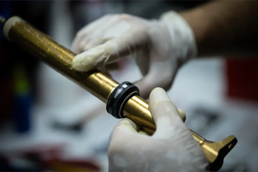 Una persona que lleva guantes coloca un anillo negro en un tubo metálico de color dorado, que podría formar parte de un componente mecánico o de automoción.