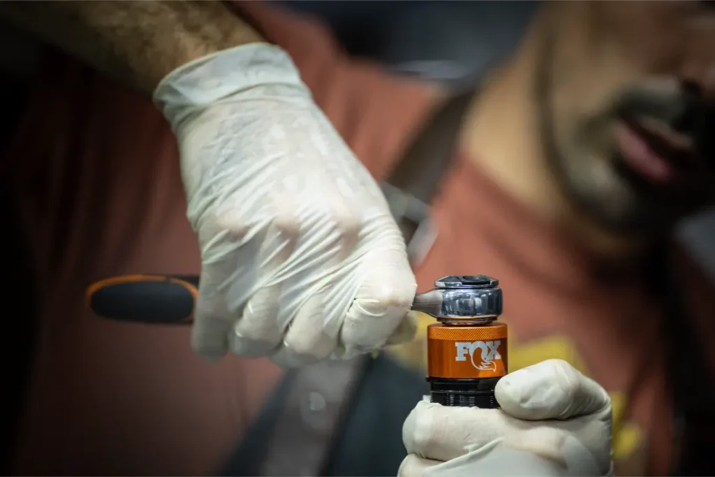 Una persona que lleva guantes utiliza una herramienta para ajustar un amortiguador FOX de color naranja.