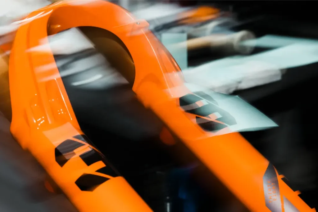 Primer plano de una horquilla de suspensión naranja para bicicleta de montaña con motivos gráficos negros, fotografiada en un interior con el fondo desenfocado.