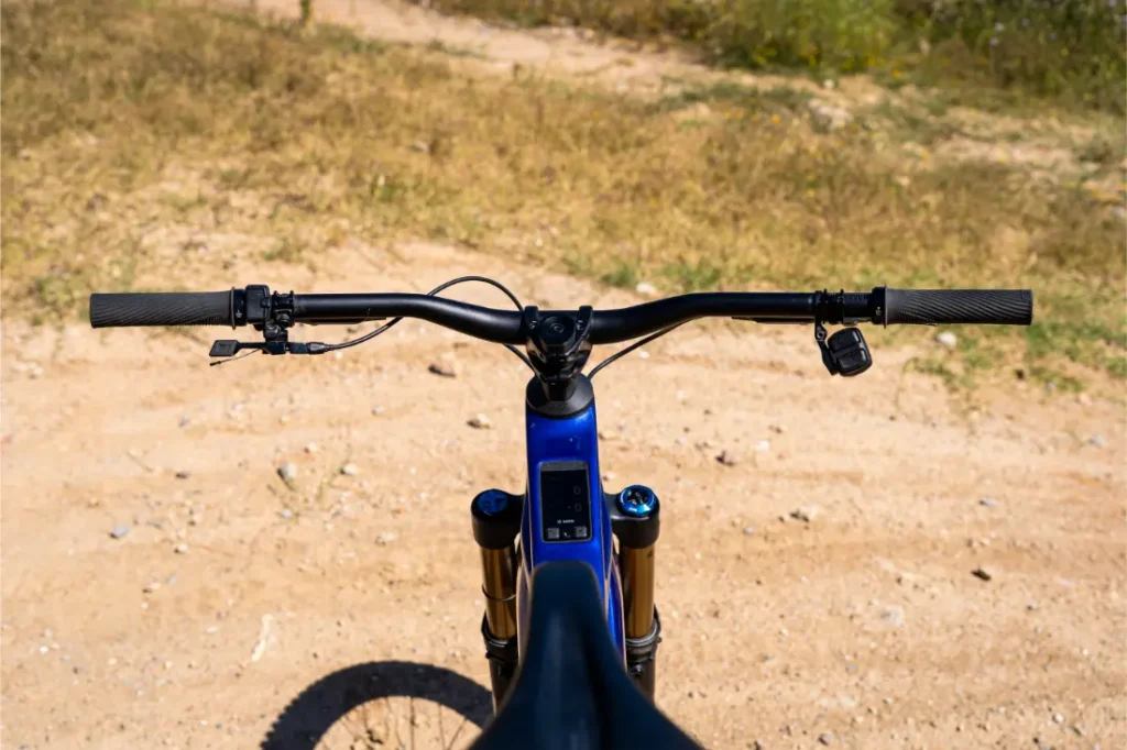 Una toma desde el punto de vista del manillar y la rueda delantera de una bicicleta de montaña azul en un sendero de tierra, rodeada de hierba seca, transmite la sensación de control y potencia potencial, ideal para los ciclistas que buscan la actualización de rendimiento de Bosch.