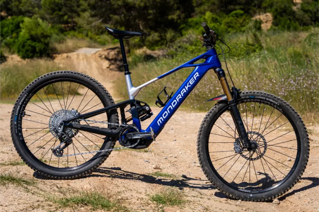 Una bicicleta de montaña Mondraker azul y blanca, que destaca por su impresionante control, está aparcada en un sendero de tierra, rodeada de vegetación herbácea y árboles al fondo.