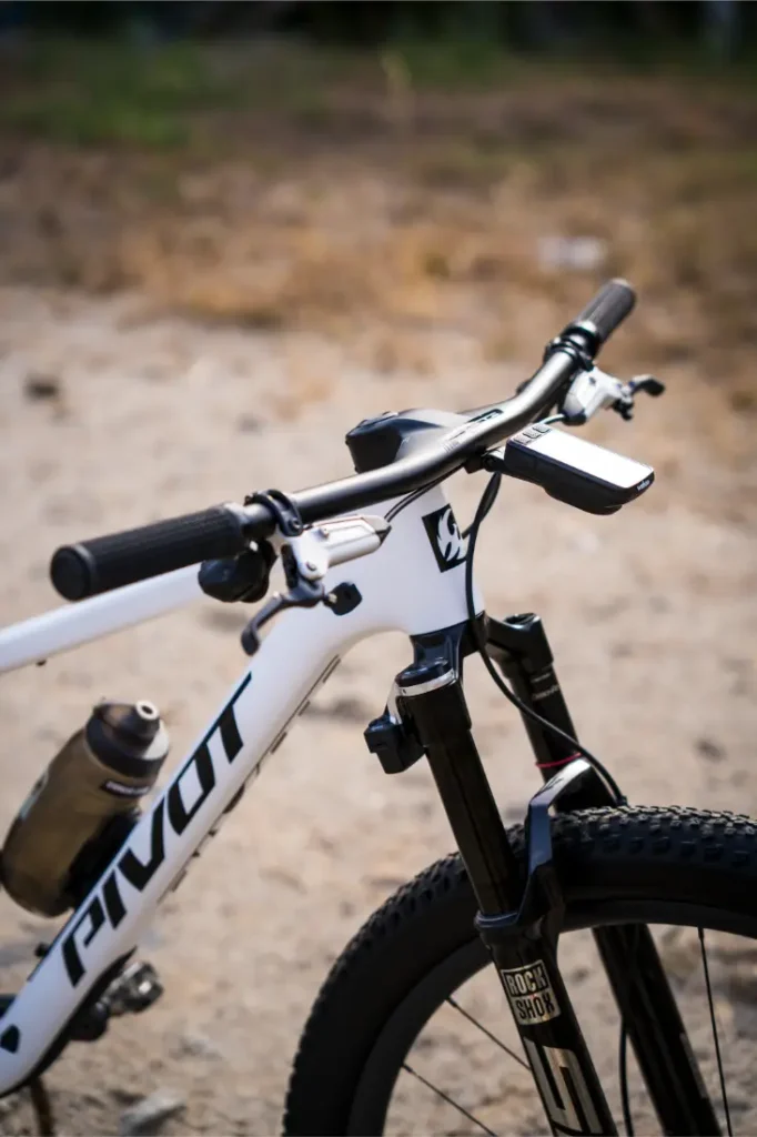 Primer plano del manillar y la suspensión delantera de una bicicleta de montaña Pivot blanca, con un manillar integrado de carbono FSA NS SIC y un portabotellas montado en el cuadro, al aire libre sobre una superficie de tierra.
