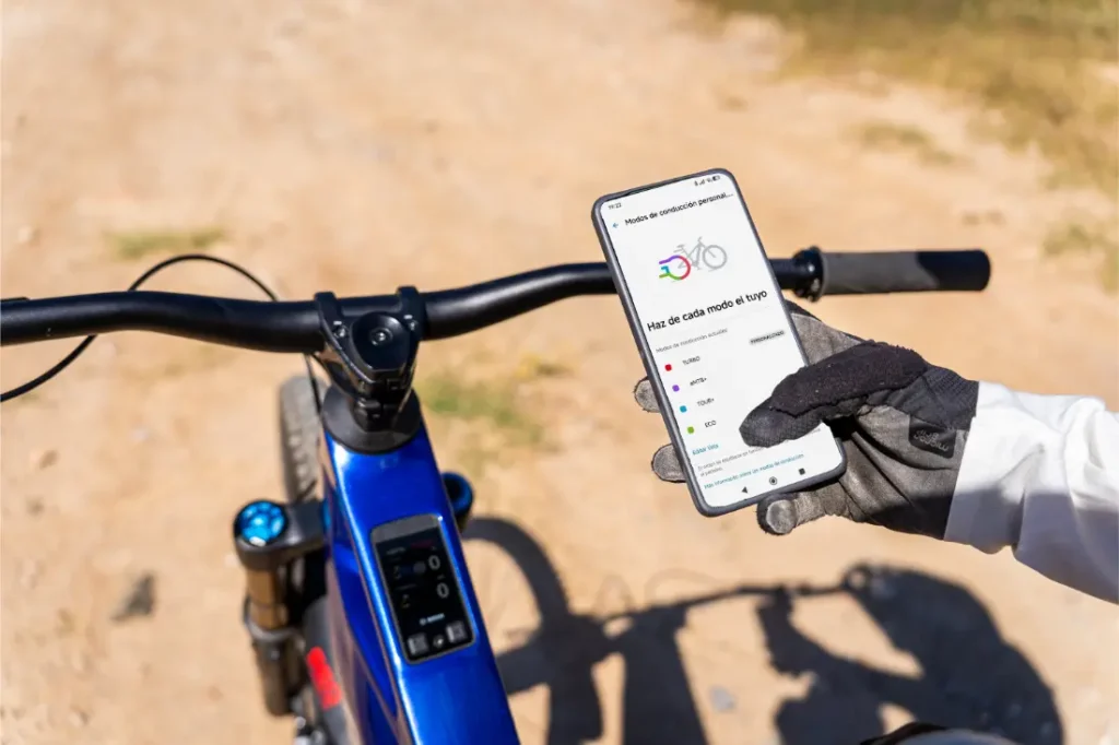 Una persona con guantes sostiene un smartphone en el que se ve una aplicación de ciclismo, mientras aparca una bicicleta azul equipada con Bosch Performance Upgrade en un camino de tierra al aire libre, disfrutando de más control y más potencia durante su recorrido.