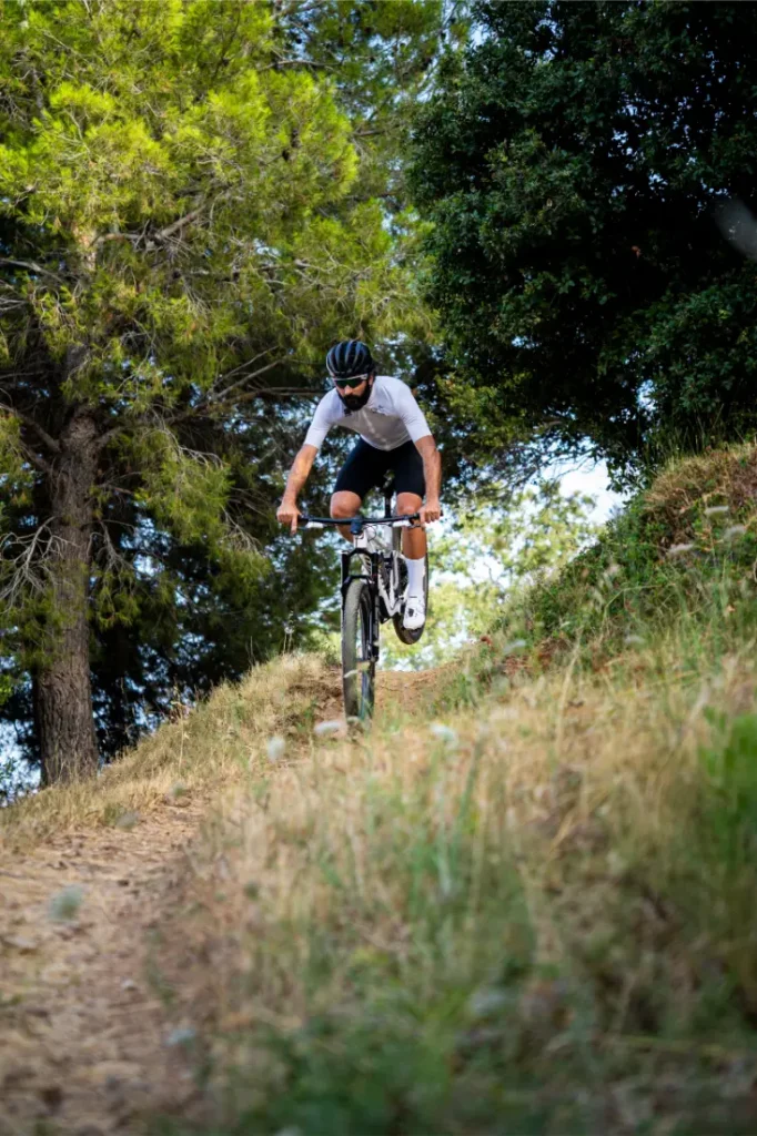 Una persona que lleva casco y gafas de sol recorre en bicicleta de montaña, equipada con un manillar integrado de carbono FSA NS SIC, un sendero de tierra rodeado de hierba y árboles.