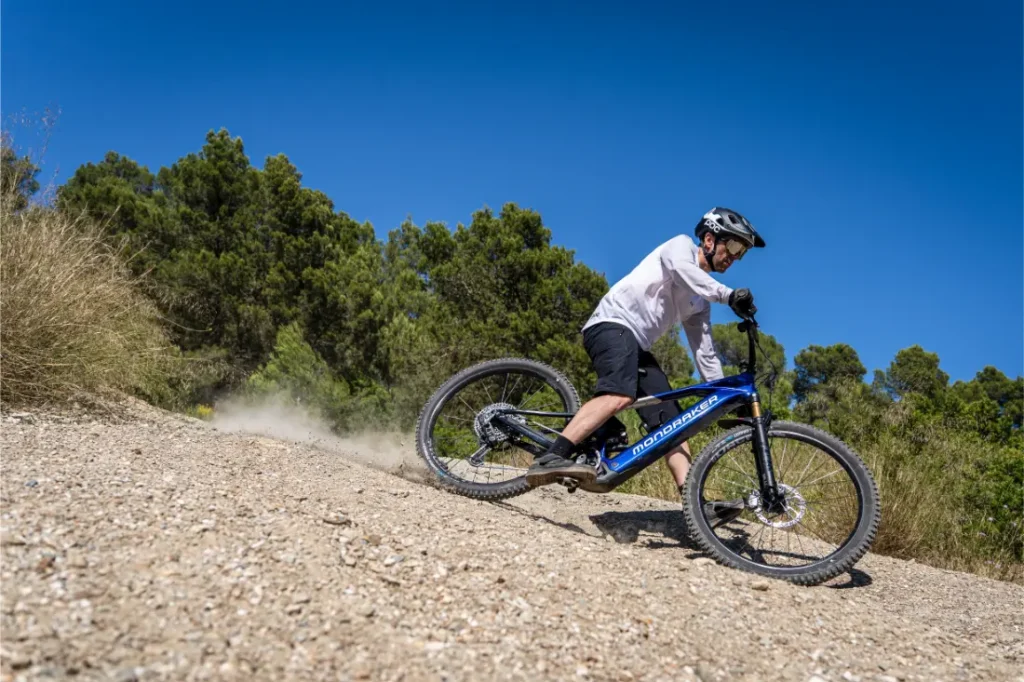 Una persona con casco baja en bicicleta de montaña azul por un sendero rocoso, rodeada de árboles y bajo un cielo azul y despejado. Gracias al Bosch Performance Upgrade, disfruta de una mayor potencia y control mientras el polvo se levanta tras la rueda trasera.