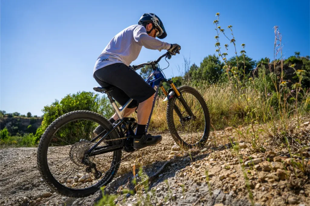 Una persona con casco sube en bicicleta de montaña por un sendero rocoso, rodeada de hierba seca y vegetación, bajo un cielo azul y despejado, disfrutando de «más potencia y más control» gracias al Bosch Performance Upgrade.