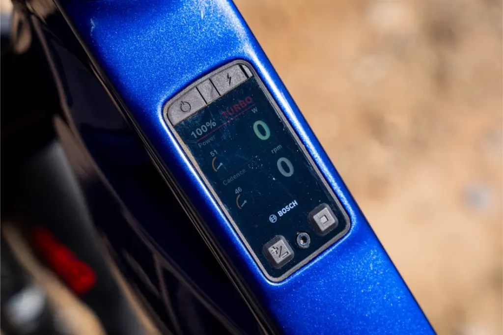 Primer plano de una pantalla digital en una bicicleta eléctrica azul, en la que se muestran el 100 % de la batería, los ajustes de potencia, la cadencia, la velocidad y la distancia recorrida, con la marca Bosch visible. Disfruta de un mayor control y potencia con la actualización de rendimiento de Bosch para una experiencia de conducción optimizada.
