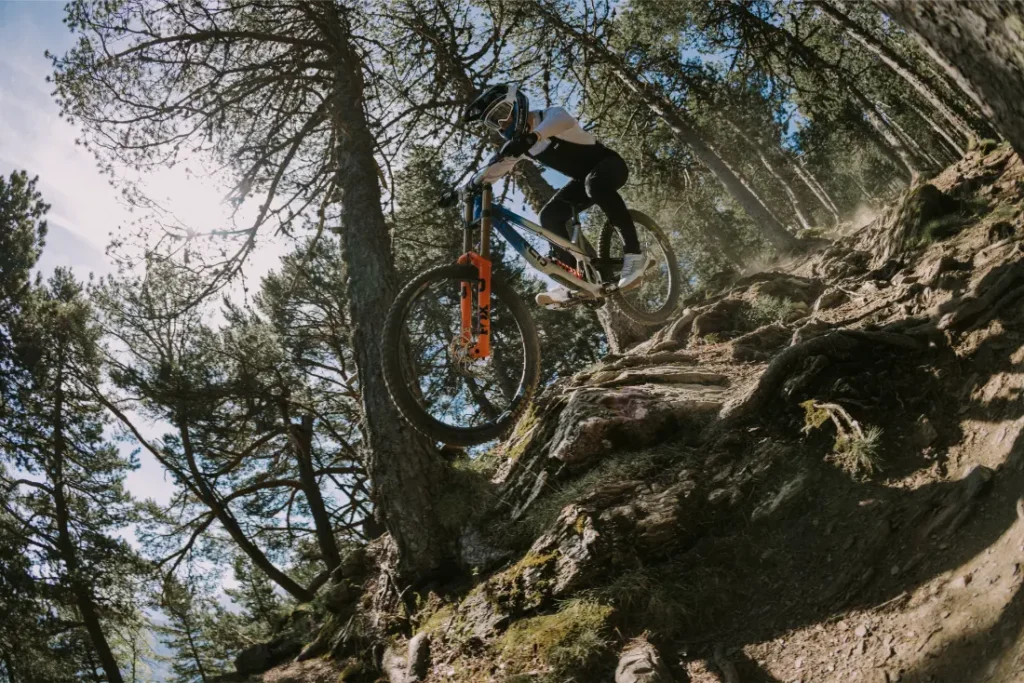 Con un casco integral POC Barocon Carbon, una persona desciende en bicicleta de montaña por un terreno irregular y cubierto de raíces en un bosque bañado por los rayos del sol.