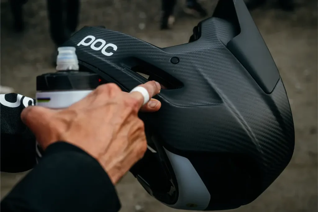 Una persona sostiene un casco integral negro POC Barocon Carbon mientras aprieta una botella de agua por encima de él, lo que transmite una sensación dinámica de descenso.