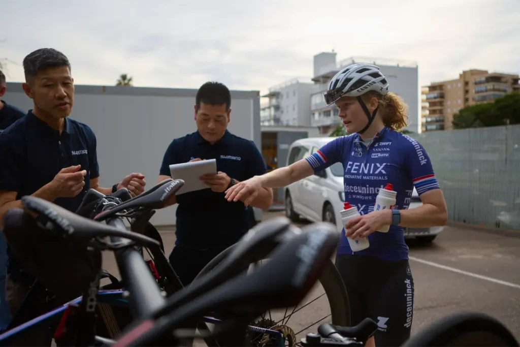 Un ciclista con casco habla con dos hombres junto a unas bicicletas aparcadas al aire libre; su atención se centra momentáneamente en un conjunto de bicicletas equipadas con ruedas Shimano Dura-Ace. Uno de los hombres escribe en un portapapeles, mientras que el otro observa. Al fondo se ven algunos edificios.