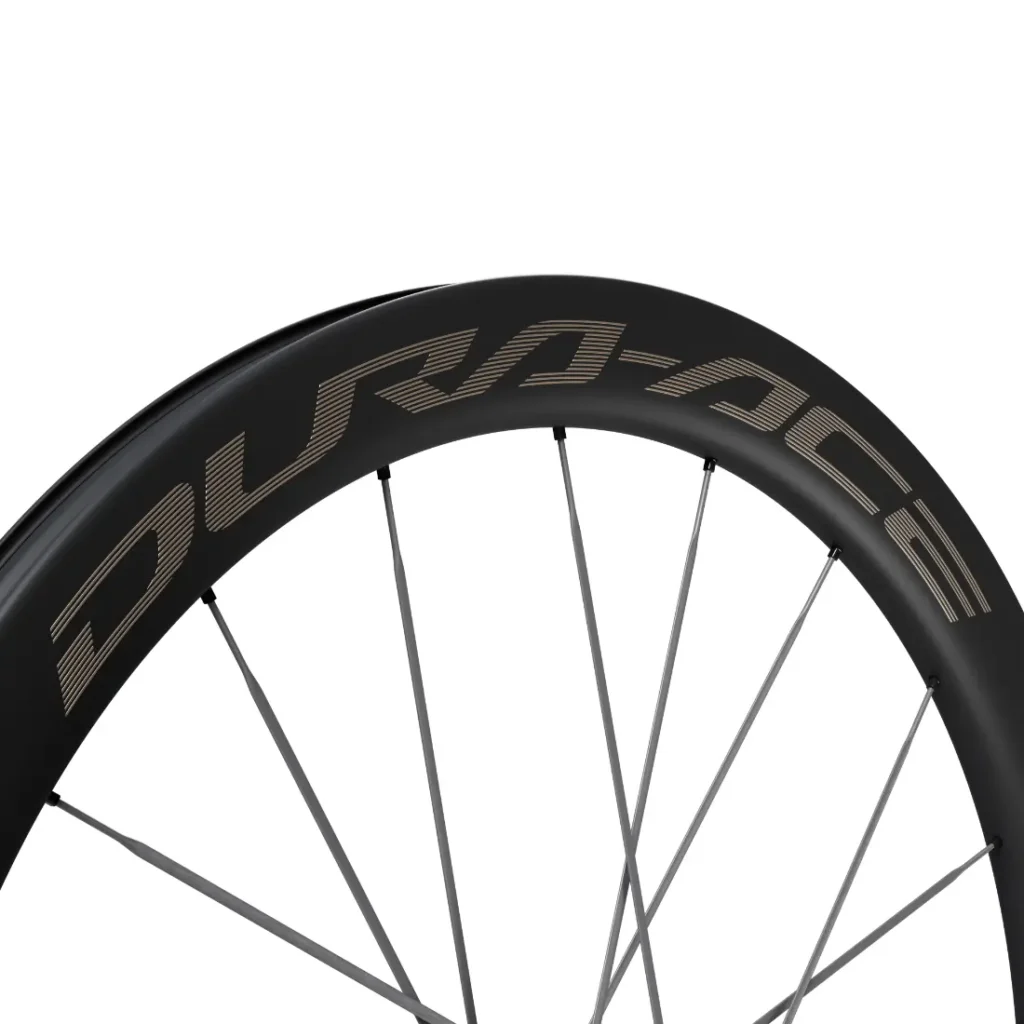 Primer plano de una llanta de bicicleta de carbono negro con la marca «DURA-ACE» en letras grandes, en la que se aprecian varios radios plateados. Este modelo Dura-Ace de Shimano, el WH-R9370, destaca por la precisión y la ligereza características de las ruedas de carbono.
