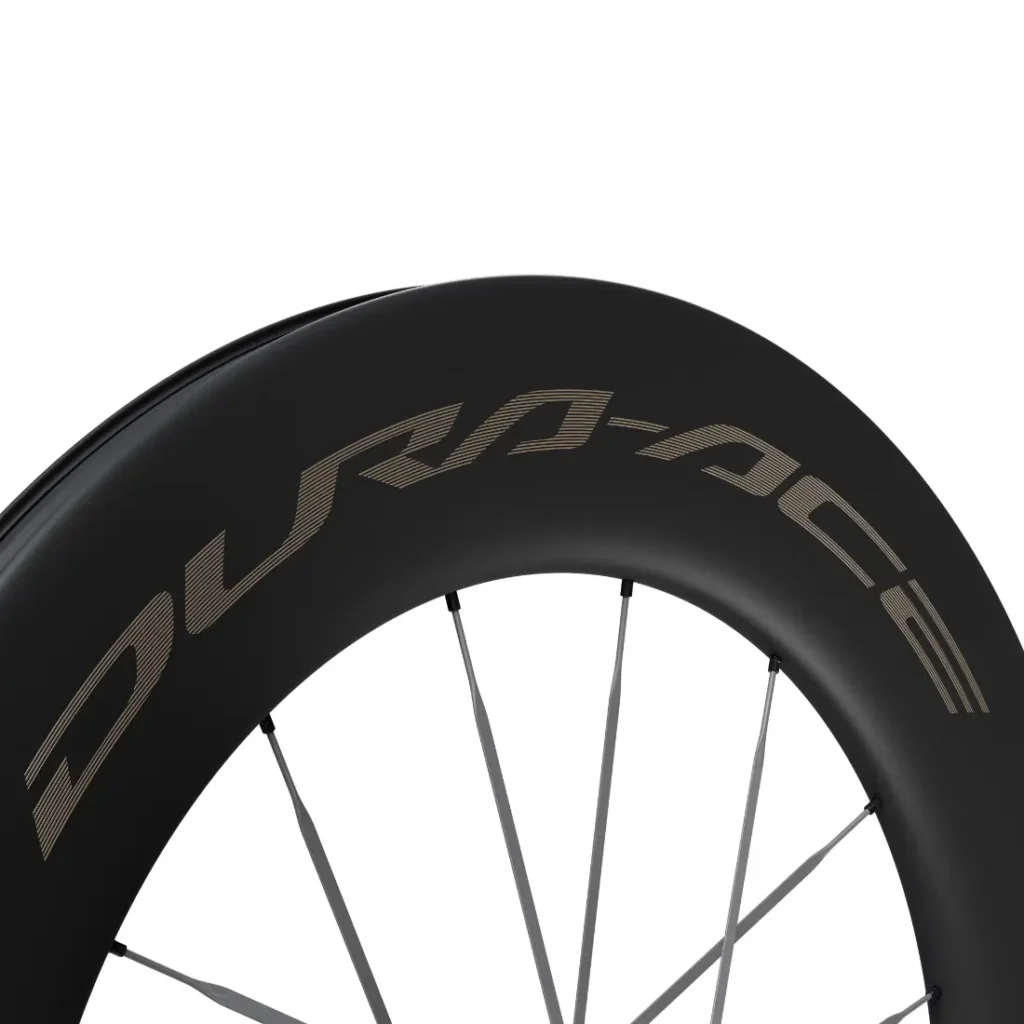 Primer plano de una rueda de bicicleta de carbono negro con radios plateados, en la que destaca la distintiva marca «DURA-ACE» en la llanta, escrita con letras grandes y estilizadas. Esta rueda Shimano Dura-Ace WH-R9370 destaca por su avanzado diseño de radios de carbono, que mejora el rendimiento y la durabilidad.