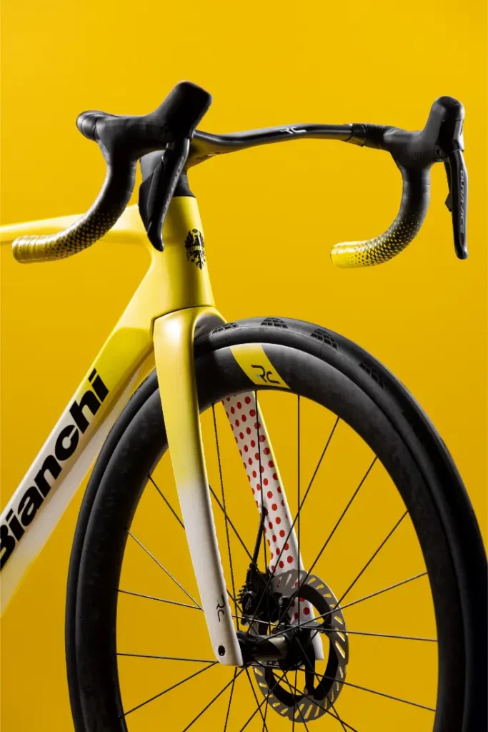 Un primer plano de la parte delantera de una bicicleta de carretera Bianchi amarilla y blanca, en el que se aprecian el manillar, la horquilla con lunares rojos, la rueda y el freno de disco, sobre un fondo amarillo.