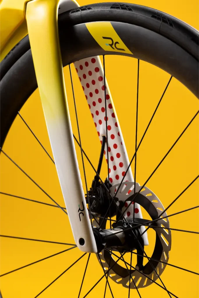 Primer plano de la rueda delantera de una bicicleta con freno de disco y horquilla amarilla, con lunares rojos en la parte interior de la horquilla, sobre un fondo amarillo.