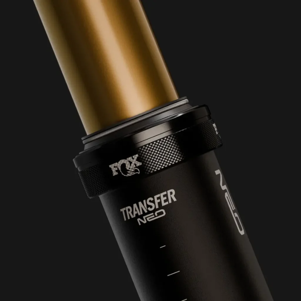 Primer plano de una tija telescópica electrónica FOX Transfer Neo E-Optimized para bicicleta de montaña, en el que se aprecia el tubo central dorado y la sección inferior negra con el logotipo sobre un fondo oscuro.