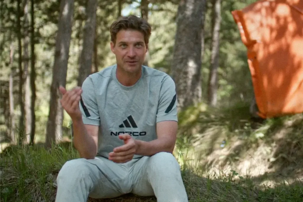 Un hombre con una camiseta gris de Adidas de la marca Norco está sentado al aire libre sobre la hierba, haciendo gestos con las manos. Cerca de él, un casco integral de la marca POC deja entrever su pasión por el descenso en bicicleta. Al fondo se ven árboles y una tienda de campaña naranja.