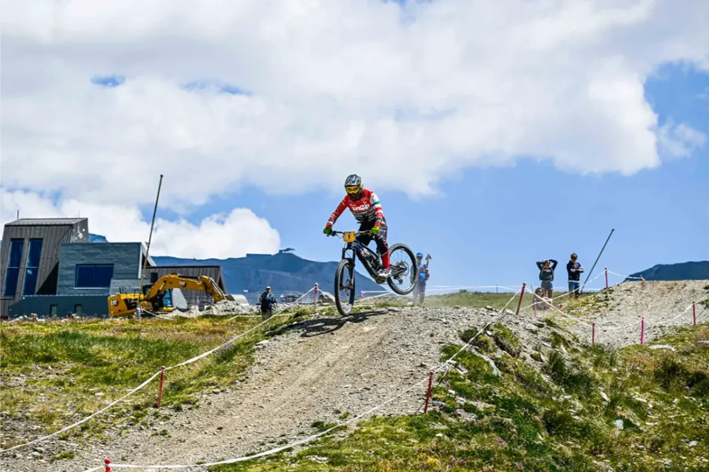 Un ciclista de montaña, con todo el equipo puesto, salta por encima de una rampa de tierra en un sendero al aire libre, mientras los espectadores observan y se ve un vehículo de obra al fondo.