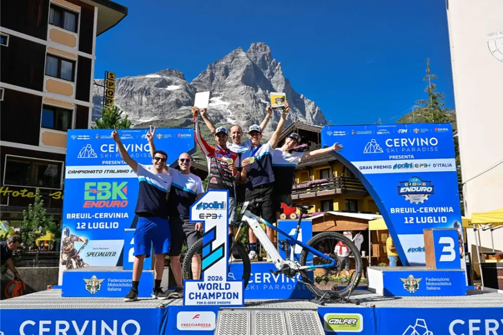 Cinco ciclistas posan en un podio con trofeos y medallas en la mano, con una montaña y pancartas del evento al fondo, en un campeonato de ciclismo.