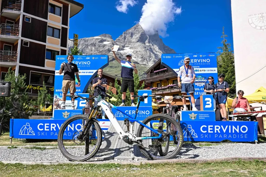 Cuatro ciclistas posan en el podio de los ganadores con una bicicleta de montaña delante, pancartas azules de «Cervino Ski Paradise» detrás de ellos y una cima de montaña al fondo, bajo un cielo despejado.