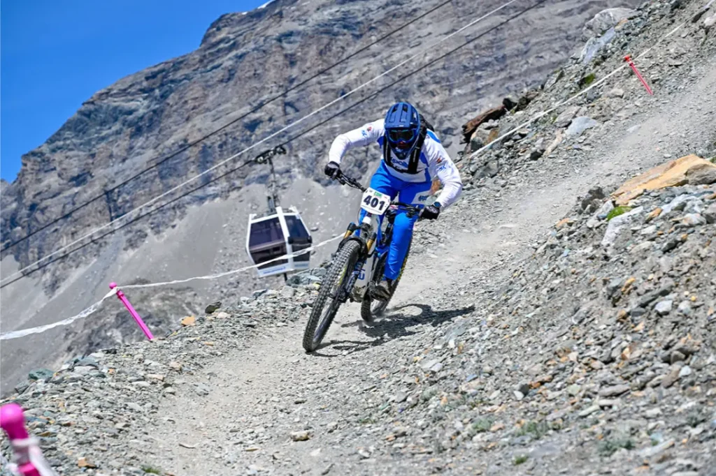 Un ciclista de montaña, vestido con ropa azul y blanca, desciende por un sendero rocoso; al fondo se ve una placa con el número 401 y un teleférico.