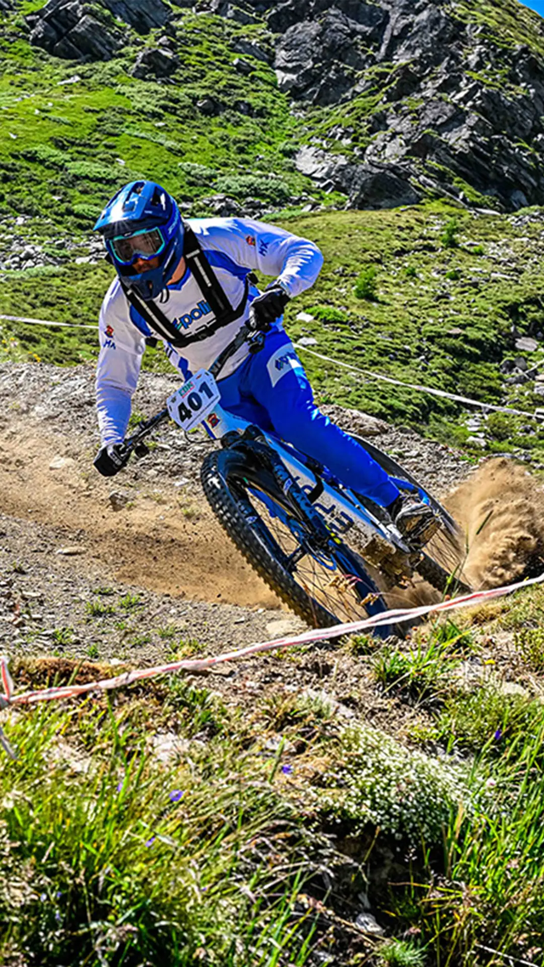 Un ciclista de montaña, vestido con un maillot azul y blanco, con casco y el dorsal 401, desciende por un sendero de tierra rodeado de hierba y terreno rocoso.