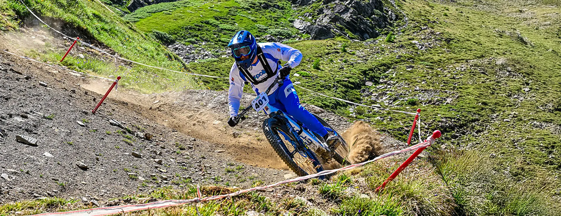 Un ciclista con equipación azul y blanca baja en bicicleta de montaña por un sendero rocoso y de tierra en la ladera cubierta de hierba, levantando una nube de polvo a su paso.