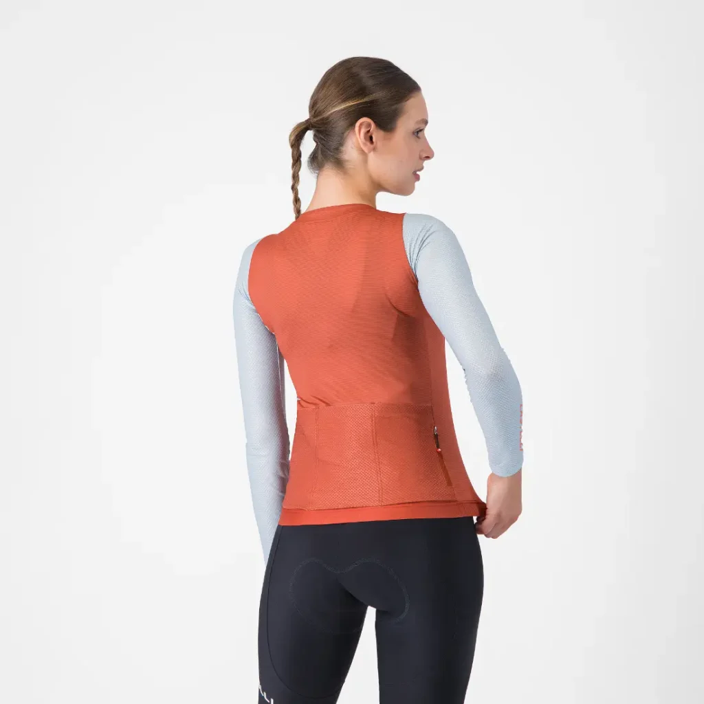Una mujer con una trenza lleva un maillot Corretto LS de manga larga y un chaleco naranja, combinados con unos culotes negros de ciclismo, y aparece de pie, de espaldas a la cámara, frente a un fondo liso.
